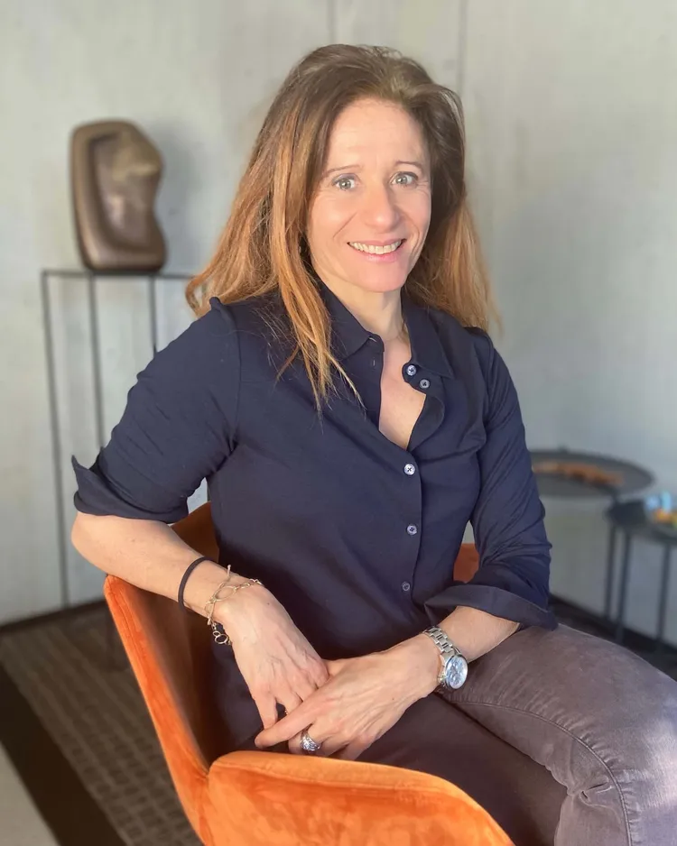 Portrait photographique d’Anne Zürcher Kondratowicz, souriante, assise dans un fauteuil orange, en chemise bleu marine, devant un mur clair.